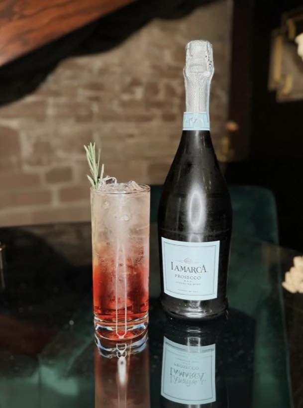 A tall cranberry highball with rosemary beside a prosecco bottle